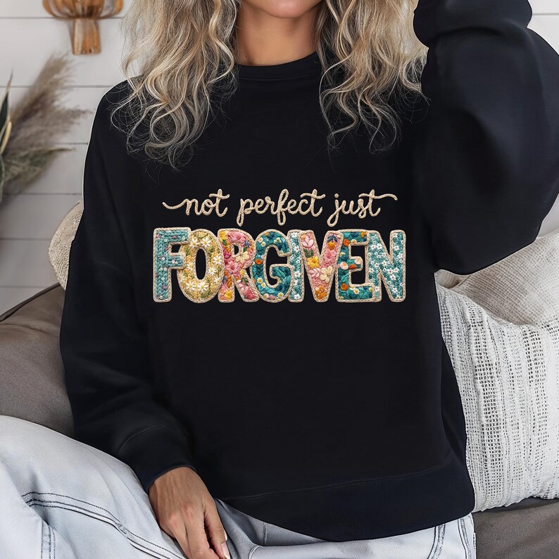 Op de afbeelding: Zwarte sweatshirt met de tekst "not perfect just FORGIVEN". Het woord "FORGIVEN" is gevuld met een bloemenpatroon in tinten geel, roze en blauwgroen. De tekst is in een scriptletter.