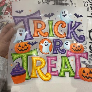 Może przedstawiać: Kolorowa grafika o tematyce Halloween z napisem "Trick or Treat" w dużych, stylizowanych literach. Projekt zawiera dynie, duchy, nietoperze i babeczkę, wszystko w żywych odcieniach pomarańczu, fioletu, zieleni i ż&oacute;łci.