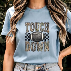 Pode incluir: Uma t-shirt azul claro com uma estampa gr&aacute;fica com o texto TOUCH DOWN num padr&atilde;o de camuflagem. Centrada entre as palavras est&aacute; uma bola de futebol americano castanho-escura com atacadores brancos sobre um fundo axadrezado preto e branco.