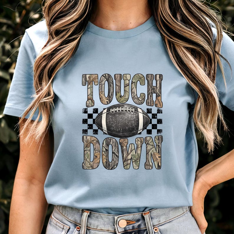 Pode incluir: Uma t-shirt azul claro com uma estampa gr&aacute;fica com o texto TOUCH DOWN num padr&atilde;o de camuflagem. Centrada entre as palavras est&aacute; uma bola de futebol americano castanho-escura com atacadores brancos sobre um fundo axadrezado preto e branco.