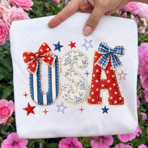 May include: A white t-shirt featuring the letters USA in large fabric applique. The U has blue and white stripes, the S has a blue floral pattern, and the A is red with white stars. Each letter is trimmed with lace and topped with a fabric bow.