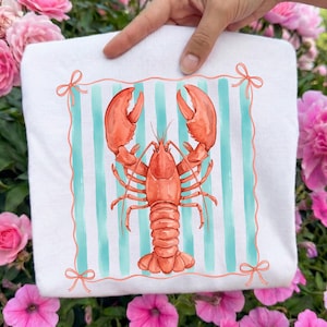 May include: White sweatshirt featuring a watercolor illustration of a red lobster. The design includes a square frame with teal stripes and coral bows. The lobster is centered on the front of the shirt.
