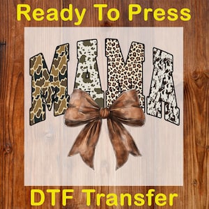 May include: A DTF transfer featuring the word "MAMA" in a mix of camouflage, cow print, and leopard print patterns. A large brown bow is centered below the word. The text "Ready To Press" and "DTF Transfer" are displayed in yellow.