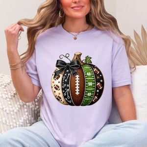 May include: A light purple short-sleeve t-shirt featuring a central graphic of a pumpkin designed with American football elements. The pumpkin includes a brown football texture, a green field with yard lines, and a black bow tied at the top stem.