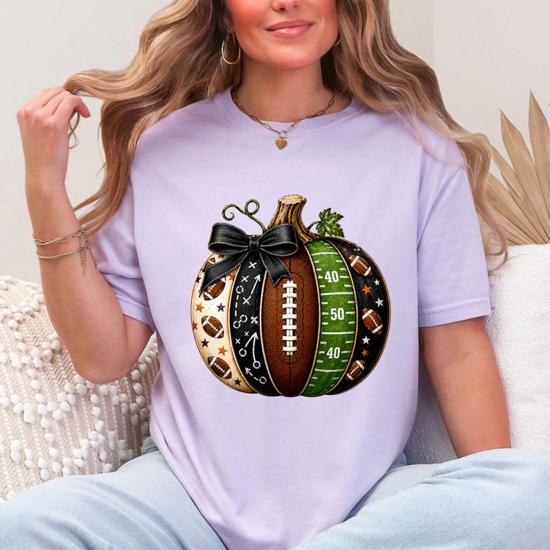 May include: A light purple short-sleeve t-shirt featuring a central graphic of a pumpkin designed with American football elements. The pumpkin includes a brown football texture, a green field with yard lines, and a black bow tied at the top stem.