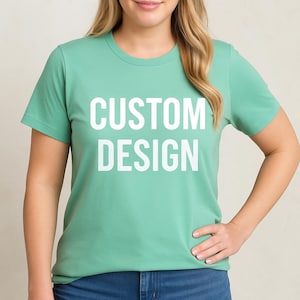May include: A light green t-shirt with the words "CUSTOM DESIGN" printed in large white letters. The shirt has short sleeves and a crew neck. The model is wearing blue jeans.