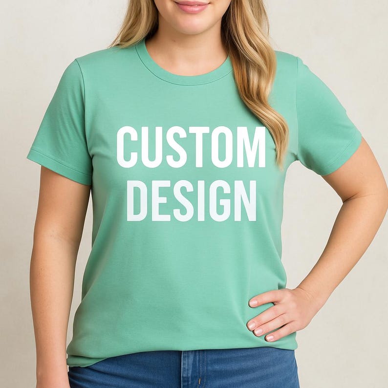 May include: A light green t-shirt with the words "CUSTOM DESIGN" printed in large white letters. The shirt has short sleeves and a crew neck. The model is wearing blue jeans.
