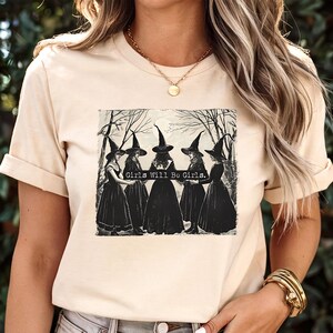 Puede incluir: Camiseta beige con un gr&aacute;fico de cinco brujas con vestidos y sombreros negros, con el texto "Girls Will Be Girls". El dise&ntilde;o es una ilustraci&oacute;n vintage en blanco y negro. La camiseta tiene mangas cortas y cuello redondo.