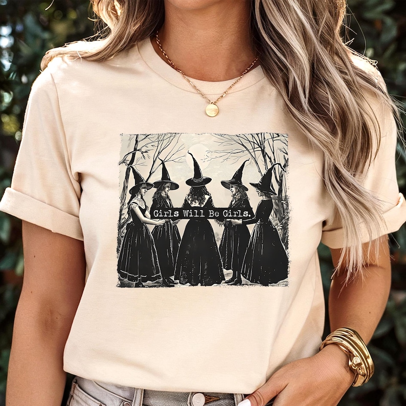 Puede incluir: Camiseta beige con un gr&aacute;fico de cinco brujas con vestidos y sombreros negros, con el texto "Girls Will Be Girls". El dise&ntilde;o es una ilustraci&oacute;n vintage en blanco y negro. La camiseta tiene mangas cortas y cuello redondo.