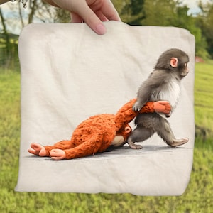 May include: A light-colored, rectangular throw blanket featuring a whimsical illustration of two monkeys. One monkey, with gray fur, is walking while holding the arm of an orange monkey lying on the ground. The background is a blurred green field.