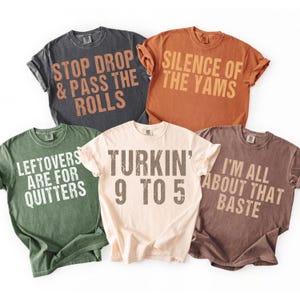 Puede incluir: Cinco camisetas de manga corta y cuello redondo en gris oscuro, naranja, verde oliva, crema y marrón. Cada camiseta presenta texto desgastado: STOP DROP & PASS THE ROLLS, SILENCE OF THE YAMS, LEFTOVERS ARE FOR QUITTERS, TURKIN' 9 TO 5, I'M ALL ABOUT THAT BASTE.