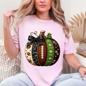 May include: A light pink t-shirt featuring a graphic of a pumpkin designed with American football elements. The pumpkin includes a black bow, football laces, a green field section with yard lines, and small football and star patterns.