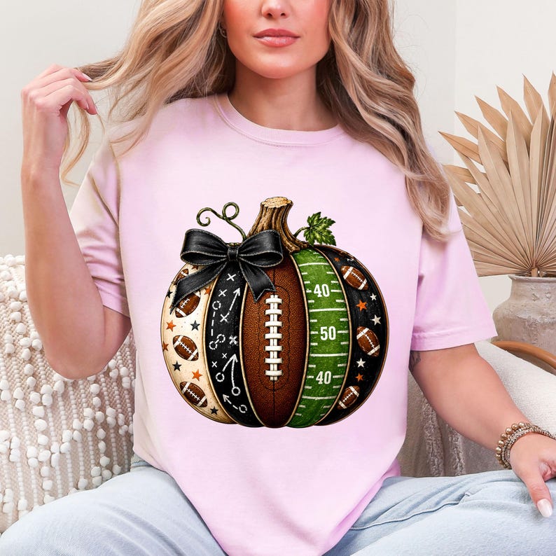 May include: A light pink t-shirt featuring a graphic of a pumpkin designed with American football elements. The pumpkin includes a black bow, football laces, a green field section with yard lines, and small football and star patterns.
