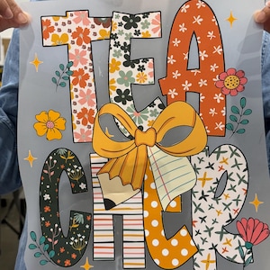 Op de afbeelding: Een kleurrijke afbeelding met het woord 'TEACHER' in grote, versierde letters. De letters zijn gevuld met bloemmotieven, stippen en strepen. Een grote gele strik en potlood staan centraal, met bloemen en sterren.