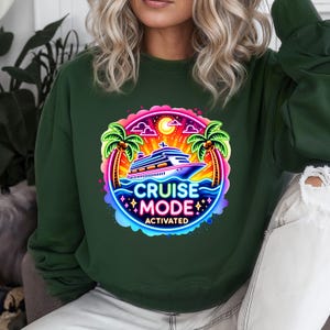 May include: Dark green sweatshirt featuring a vibrant graphic. The graphic includes a cruise ship, palm trees, and the words "CRUISE MODE ACTIVATED" in a neon-style design. The design is set against a backdrop of a sunset and ocean waves.