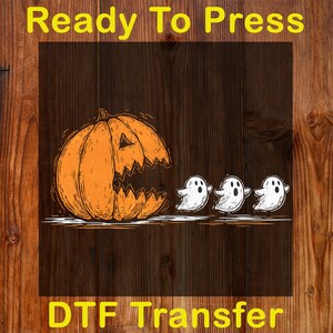 May include: Orange pumpkin with a carved face and three white ghost illustrations. The words "Ready To Press" and "DTF Transfer" are in yellow text. The background is a dark wood grain.