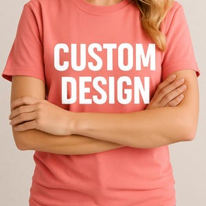 May include: A coral-colored t-shirt with the words "CUSTOM DESIGN" printed in large, white capital letters. The shirt is short-sleeved and appears to be made of a soft material. The person wearing the shirt has their arms crossed.