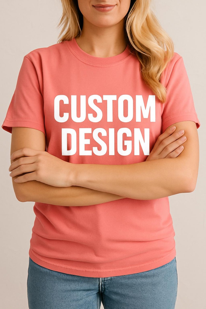May include: A coral-colored t-shirt with the words "CUSTOM DESIGN" printed in large, white capital letters. The shirt is short-sleeved and appears to be made of a soft material. The person wearing the shirt has their arms crossed.