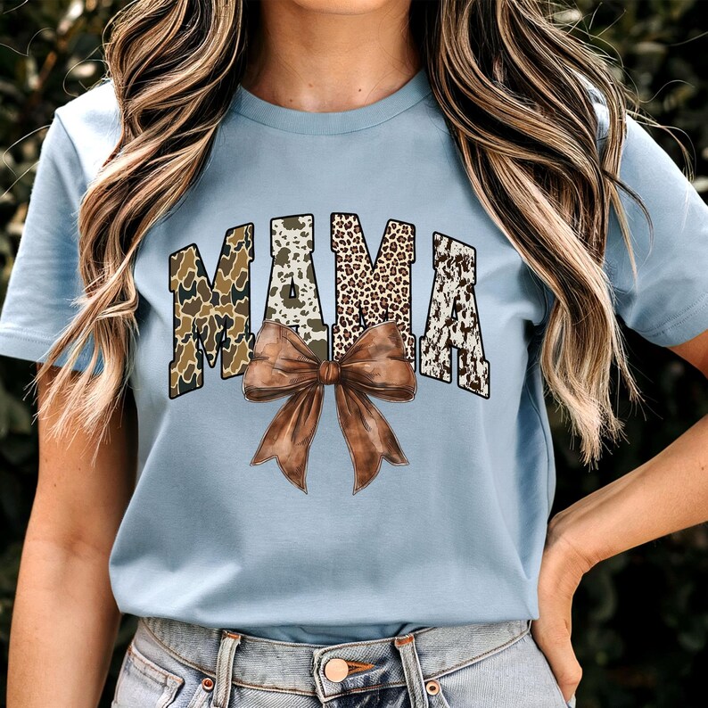May include: Light blue t-shirt featuring the word "MAMA" in a bold, patterned design. The letters have a camouflage, leopard print, and cow print pattern. A large brown bow is centered below the word, adding a decorative touch. The shirt is a casual wear item.
