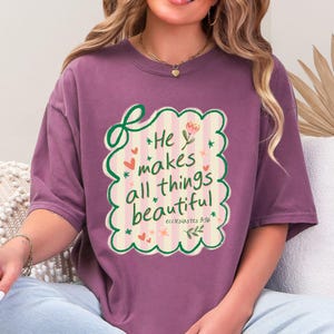 May include: Mauve-colored t-shirt with a decorative design. The design features the text "He makes all things beautiful" in a whimsical font, surrounded by hearts, flowers, and a scalloped border. The text is from Ecclesiastes 3:11.