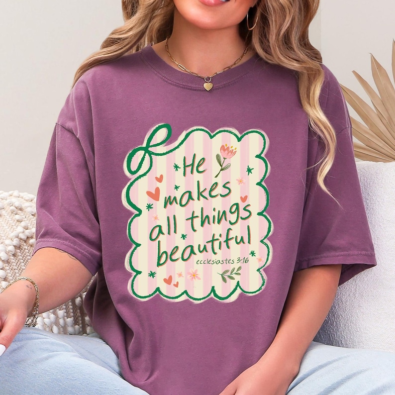 May include: Mauve-colored t-shirt with a decorative design. The design features the text "He makes all things beautiful" in a whimsical font, surrounded by hearts, flowers, and a scalloped border. The text is from Ecclesiastes 3:11.