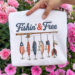 May include: White sweatshirt with the text "Fishin' & Free" in navy and red. Below the text is a fishing rod with six fishing lures hanging from it. The lures have red, white, and blue designs.