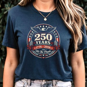 Peut inclure: T-shirt bleu marine avec un motif circulaire comm&eacute;morant 250 ans. Le design comprend un aigle, des &eacute;toiles et le texte "250 YEARS 1776-2026 We The People" en rouge, blanc et bleu. Un collier dor&eacute; est visible.