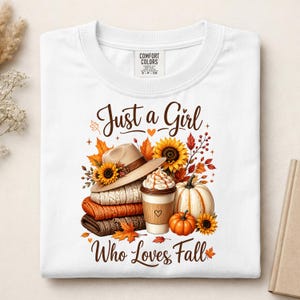 May include: A white t-shirt featuring a graphic print with the text "Just a Girl Who Loves Fall". The design includes a tan hat, stacked sweaters, a coffee cup with whipped cream, pumpkins, sunflowers, and autumn leaves in shades of orange and brown.