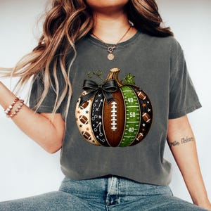 May include: A charcoal gray t-shirt featuring a graphic of a pumpkin designed with American football elements. The pumpkin includes sections with football laces, a green field with yard lines, play diagrams, and small footballs, topped with a black bow.