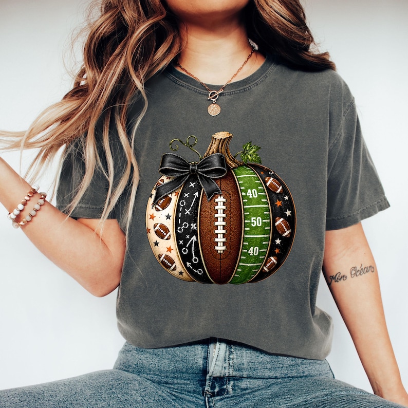 May include: A charcoal gray t-shirt featuring a graphic of a pumpkin designed with American football elements. The pumpkin includes sections with football laces, a green field with yard lines, play diagrams, and small footballs, topped with a black bow.
