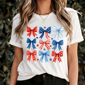 May include: White t-shirt featuring a grid of watercolor bows in red, white, and blue. The bows have star and stripe patterns, with a few blue stars scattered around. The shirt is a casual, patriotic design.