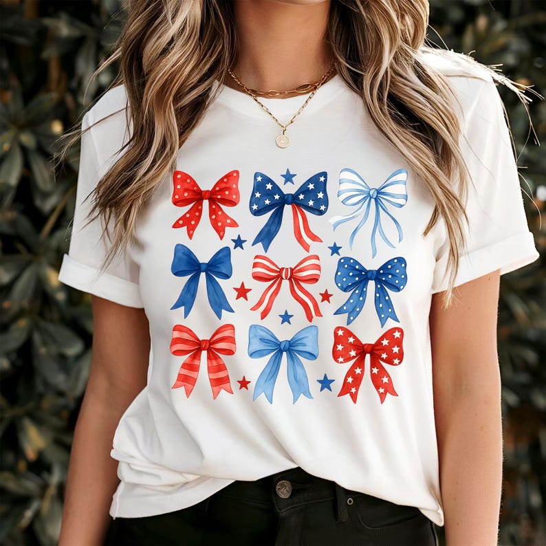 May include: White t-shirt featuring a grid of watercolor bows in red, white, and blue. The bows have star and stripe patterns, with a few blue stars scattered around. The shirt is a casual, patriotic design.