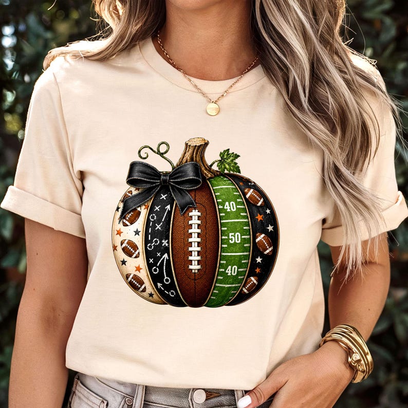 May include: A cream-colored t-shirt featuring a graphic of a pumpkin designed with American football elements. The pumpkin includes a brown football texture, a green field section with yard lines, a black bow, and small football icons.