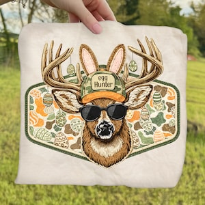 May include: A beige graphic tee featuring a deer with antlers, sunglasses, and a camouflage hat that says "Egg Hunter." The deer has bunny ears and is surrounded by Easter eggs. The design is set within a green and orange patterned border.