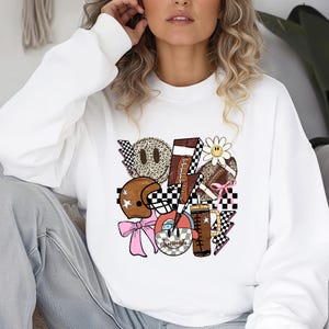 Peut inclure: Sweat-shirt blanc avec un motif graphique color&eacute;. Le motif comprend un ballon de football am&eacute;ricain, un casque, des visages souriants et des &eacute;clairs. Le graphisme int&egrave;gre des motifs tels que l'imprim&eacute; l&eacute;opard et les drapeaux &agrave; damiers, avec des accents de rose, de marron et de noir.