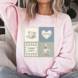 May include: A light pink sweatshirt with a patchwork design featuring a teacup, a heart with the word "Thrifters", a goose, and the text "Gonna Thrift". The design is in shades of blue, green, and white.