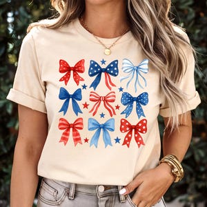 May include: A beige t-shirt featuring a pattern of red and blue bows with stars. The bows have various designs, including polka dots and stripes. The shirt has short sleeves and a crew neck.