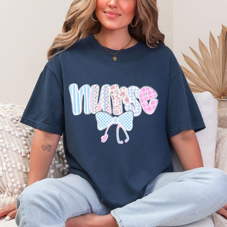 May include: Navy blue t-shirt with the word "NURSE" in a patchwork design. The letters are in various patterns, including stripes, gingham, and floral prints. A bow and stethoscope graphic is below the word.