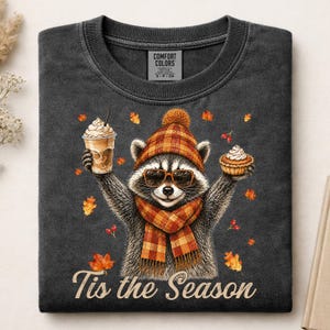 May include: A charcoal gray t-shirt featuring a graphic of a raccoon wearing a plaid hat, scarf, and sunglasses. The raccoon holds a coffee cup and a cupcake. Scattered autumn leaves and berries surround the raccoon, with the text Tis the Season.