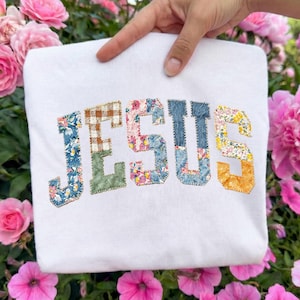 May include: A white sweatshirt with the word "JESUS" appliqued across the front. The letters are made of various floral and patterned fabrics, including blue, pink, and yellow. The sweatshirt is held up by a hand, with pink flowers in the background.