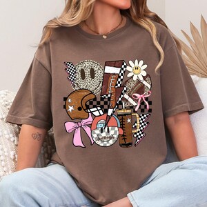 Peut inclure: T-shirt marron avec un collage de graphiques sur le th&egrave;me du football am&eacute;ricain. Le design comprend un casque, des ballons de football, une marguerite, une tasse &agrave; caf&eacute; et des &eacute;clairs, le tout avec une esth&eacute;tique r&eacute;tro et vintage. Le t-shirt a une coupe d&eacute;contract&eacute;e.