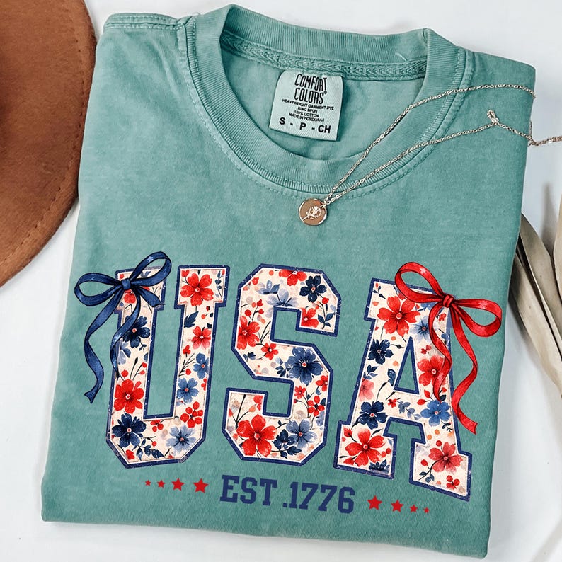 Może przedstawiać: Turkusowa koszulka Comfort Colors z napisem "USA" w kwiatowy wz&oacute;r, czerwonymi i niebieskimi kokardkami oraz napisem "EST. 1776". Srebrny naszyjnik z wisiorkiem jest zarzucony na koszulkę.