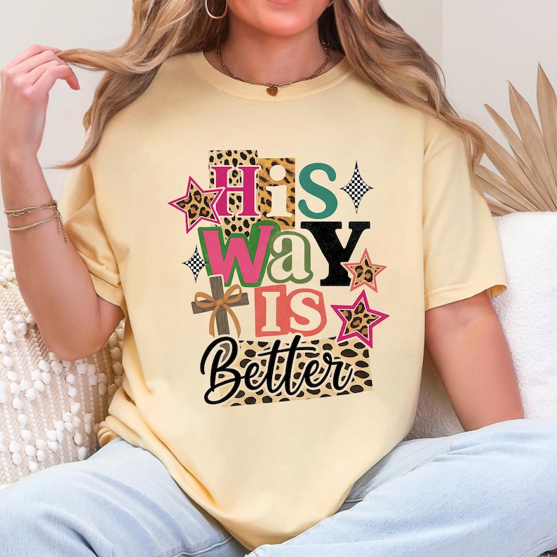 Op de afbeelding: Lichtgele t-shirt met de tekst "His Way Is Better" in een kleurrijk, patroonontwerp. Het ontwerp bevat sterren, een kruis en luipaardprint accenten. Het shirt is een casual, comfortabele stijl.
