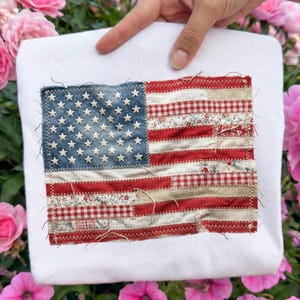 May include: White sweatshirt featuring a patchwork American flag design. The flag includes a blue field with white stars and red and white striped sections. The flag is made of various fabrics, including floral and gingham patterns.