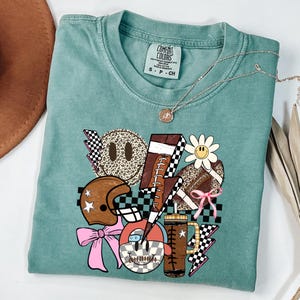 Peut inclure: Un t-shirt Comfort Colors turquoise avec un motif graphique sur le th&egrave;me du football. Le motif comprend un ballon de football, un casque, un &eacute;clair et des visages souriants avec des imprim&eacute;s l&eacute;opard et des motifs &agrave; carreaux. Un collier argent&eacute; est drap&eacute; sur le t-shirt.