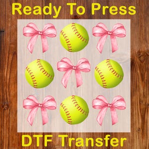 May include: A DTF transfer sheet featuring a pattern of softballs and pink bows. The softballs are yellow with red stitching, and the bows are a watercolor style. The text "Ready To Press" and "DTF Transfer" are in yellow.