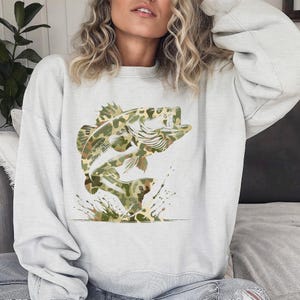 Puede incluir: Una sudadera gris claro con un dise&ntilde;o de un pez lubina saltando. El pez est&aacute; representado en un patr&oacute;n de camuflaje verde, marr&oacute;n y beige. Salpicaduras de agua se muestran debajo del pez, agregando un elemento din&aacute;mico al dise&ntilde;o.