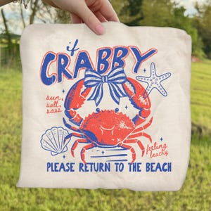 May include: Off-white tote bag with a red and blue crab graphic. The text reads "If Crabby" above the crab, and "Please Return to the Beach" below. Additional text includes "sun, salt, sass" and "feeling beachy".