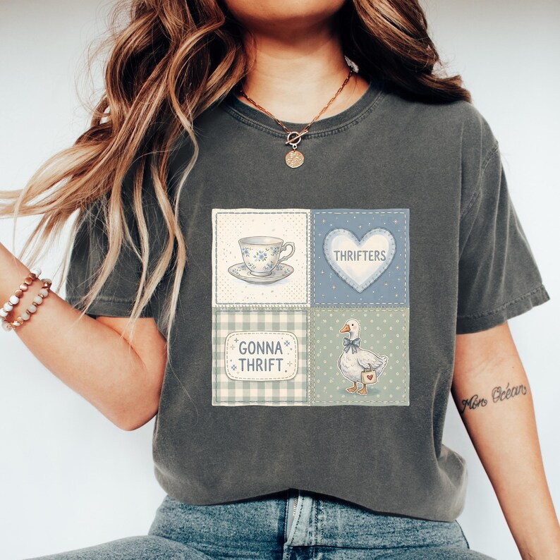 May include: Dark grey t-shirt with a patchwork design featuring a teacup, heart with "Thrifters" text, a goose, and the text "Gonna Thrift." The design incorporates blue, white, and green colors.