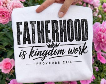 Pode incluir: Uma camiseta branca com o texto "FATHERHOOD is kingdom work" em preto, com um gráfico de coroa. O texto "PROVÉRBIOS 22:6" está abaixo. A camiseta é segurada por uma mão, rodeada de flores cor-de-rosa.
